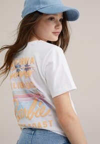 Camiseta blanca de manga corta con un gráfico multicolor que dice "Verano en California" con palmeras y el logo de "Barbie". Lleva una gorra de mezclilla azul.