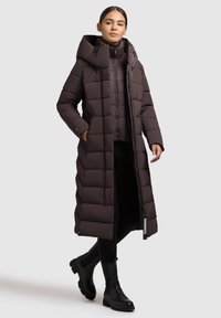 Cappotto lungo imbottito marrone con cappuccio, dotato di chiusura con zip, tasche laterali e etichetta. Indossato su un top nero e abbinato a stivali neri alla caviglia.