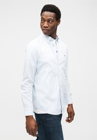 Πουκάμισο με κουμπιά στις ρίγες ανοιχτό γαλάζιο και λευκό, με γιακά, μακριά μανίκια και μικρό λογότυπο στο στήθος. Φορεμένο με σκουρόχρωμο τζιν.