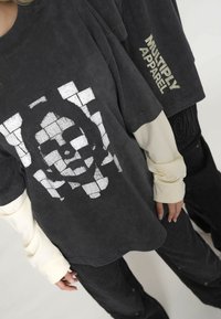 Przesadnie duża czarna koszulka z białym graficznym wzorem i beżowymi akcentami na rękawach; zawiera tekst "Multiply Apparel" w metalicznym złotym kolorze.
