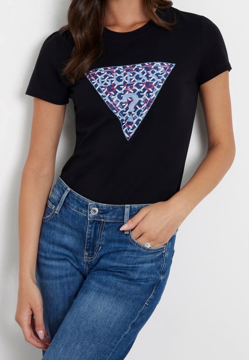 Femme portant un t-shirt noir à manches courtes avec un logo triangulaire à motifs bleu, rouge et blanc et un jean bleu avec une main dans la poche.