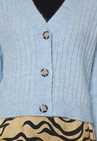 Vaaleansininen ribbineulosneuletakki V-pääntiellä, jossa on kolme tummaa nappia. Kangas vaikuttaa pehmeältä ja hieman pörröiseltä, mikä korostaa sen rakennetta.