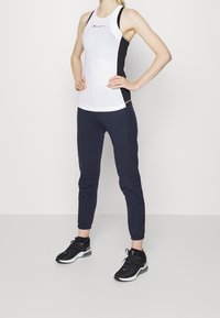 Vit sporttopp med svarta sidpaneler, i kombination med mörkblå joggers. Modellen har på sig svarta träningsskor med vita sulor.