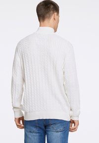 Weißer Kabelstrickpullover mit gerippten Bündchen und Saum, der vertikale strukturierte Muster und einen hohen Kragen aufweist, kombiniert mit blauen Jeans.