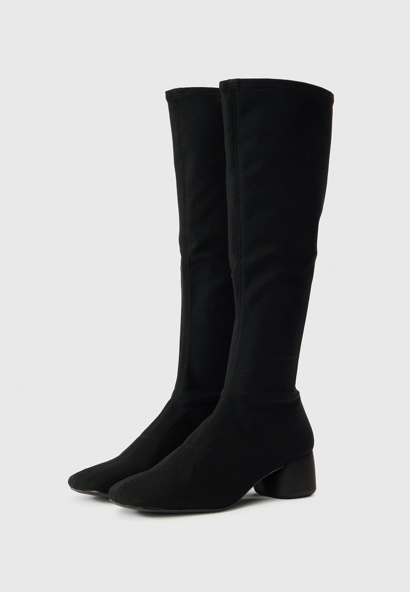 Schwarze kniehohe Stiefel aus dehnbarem Stoff mit spitz zulaufender Schuhspitze und niedrigem Blockabsatz. Nahtdetails verleihen optisches Interesse.