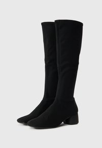 Μαύρες μπότες μέχρι το γόνατο από ελαστικό ύφασμα με μυτερή μύτη και χαμηλό χοντρό τακούνι. Οι ραφές προσθέτουν οπτικό ενδιαφέρον.