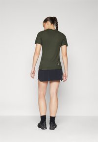 Camiseta ajustada de color verde oliva con mangas cortas, aberturas laterales en el dobladillo y una falda negra. Lleva calcetines negros y zapatillas deportivas.