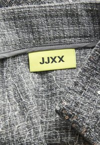 Pilkas tekstūruotas audinys su baltais siūlais ir išsibarsčiusiais blizgučiais. Etiketė su užrašu "JJXX" juodu ant geltono fono.
