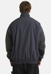 Dark gray jacket with a high collar, elastic waistband, and long sleeves. Features contrasting textured paneling and blue stitching details.