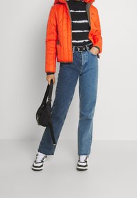 Osoba v oranžovém prošívaném kabátu, tričku s černobílým tie-dye vzorem, modrých džínách, černém opasku, bílých teniskách a držící malou černou kabelku přes rameno.