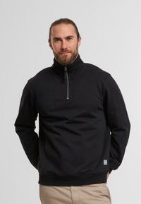 Felpa nera con zip a quarto, con collo alto, realizzata in un tessuto liscio. Presenta polsini a coste e una piccola etichetta con il logo vicino all'orlo.