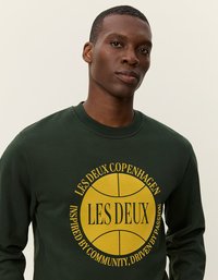 Κοντό μανίκι σκούρο πράσινο φούτερ από βαμβάκι, με κυκλική κίτρινη γραφική παράσταση και κείμενο. Ριμπαριστό γιακά και μανσέτες, μαλακή υφή.