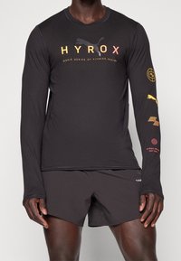 Maglietta atletica nera a maniche lunghe con grafiche del logo giallo e rosso, abbinata a pantaloni corti neri. Design leggero e aderente con cuciture piatte.