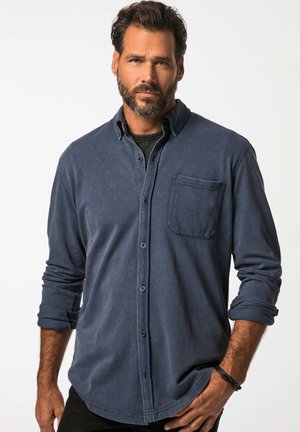 Camisa abotonada de color azul marino en una tela suave, con un bolsillo frontal, mangas dobladas y un cuello clásico; llevada con una camiseta negra debajo.