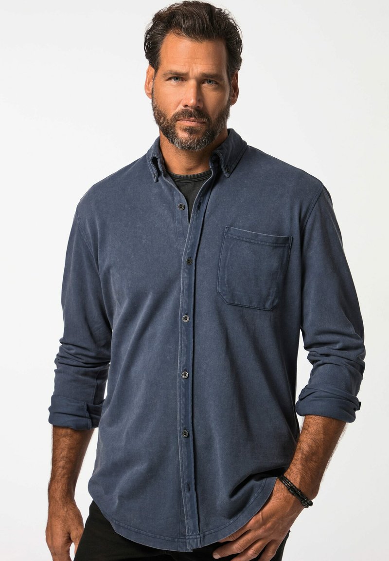 Camicia button-up blu navy in tessuto morbido con taschino anteriore, maniche arrotolate e colletto classico; indossata con una maglietta nera sotto.