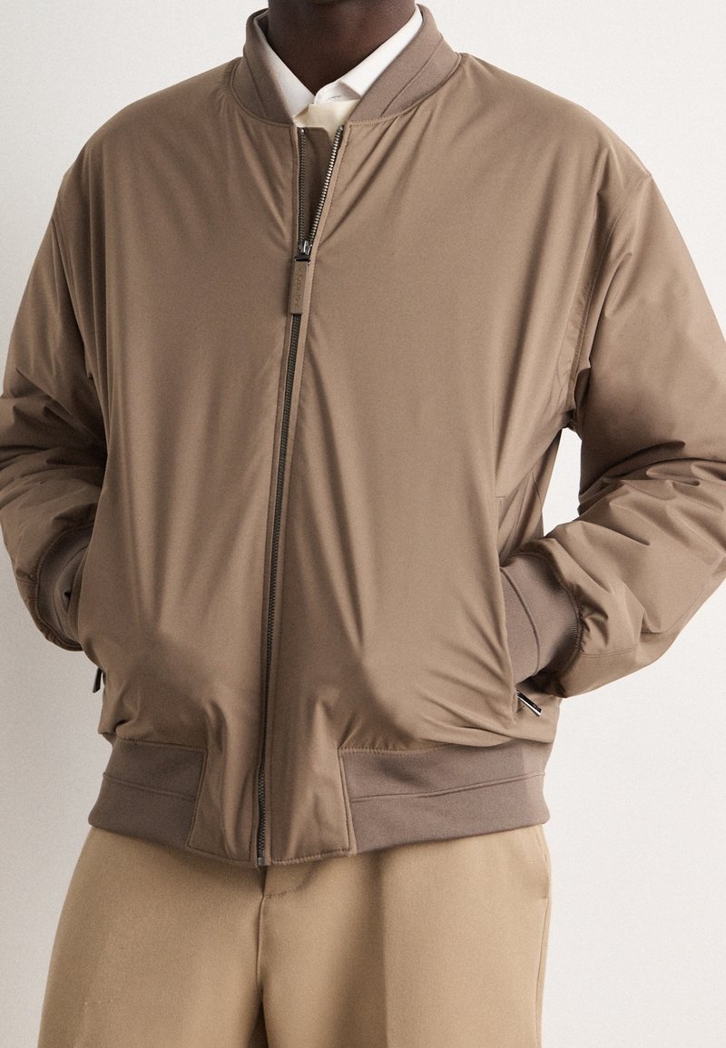 Homme portant une veste bomber marron clair zippée avec poignets et ceinture côtelés, associée à un pantalon beige et une chemise blanche à col.
