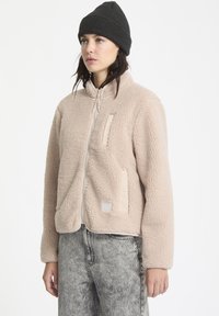 Beige fleece jack met een rits aan de voorkant, twee zijzakken en een gestructureerde stof. Gecombineerd met een zwarte gebreide beanie en grijze jeans.