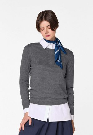 Frau trägt einen grauen Pullover über einem weißen Hemd mit Kragen, einen marineblauen Rock und ein blaues Tupftuch, das um ihren Hals gebunden ist, und blickt nach unten.