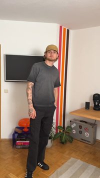 Grijze katoenen t-shirt met korte mouwen, zwarte cargo broek en zwarte sneakers. De achtergrond heeft een kleurrijke gestreepte muur en kamerplanten.