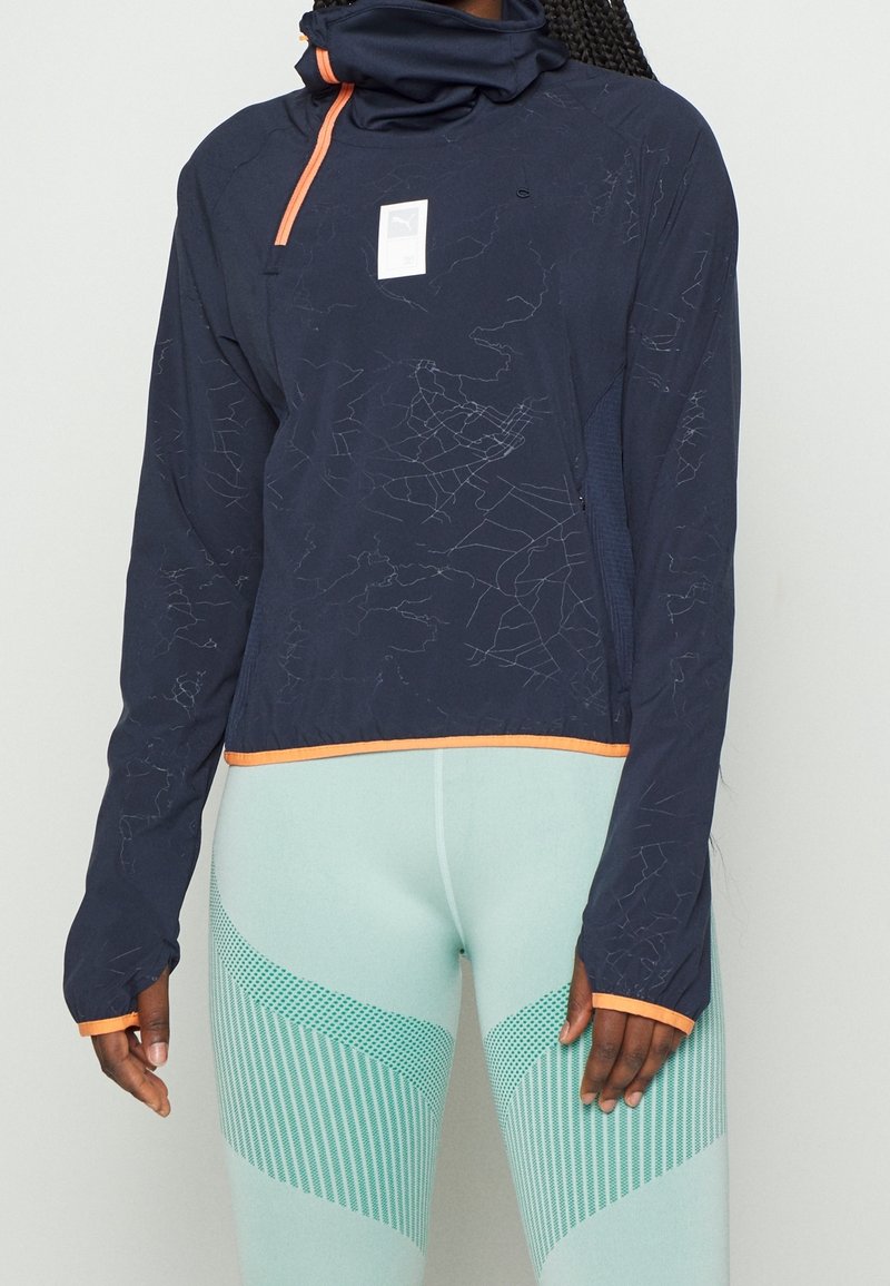 Personne portant une veste de sport bleu marine à manches longues avec de fines lignes blanches et une bordure orange, associée à des leggings texturés bleu clair.