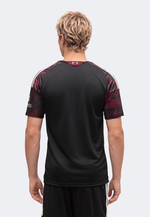 Personne blonde de dos portant un maillot de sport noir avec des manches à motif rouge foncé et un short noir à rayures blanches.