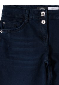 Tmavě modré džíny z denimu s hladkou texturou, které mají dva stříbrné knoflíky v pase, přední kapsu a akcentovanou nitě podél švů.