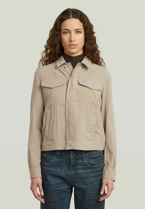 Femme aux longs cheveux bouclés portant une veste beige boutonnée sur un haut noir et un jean bleu foncé, debout devant un fond uni.