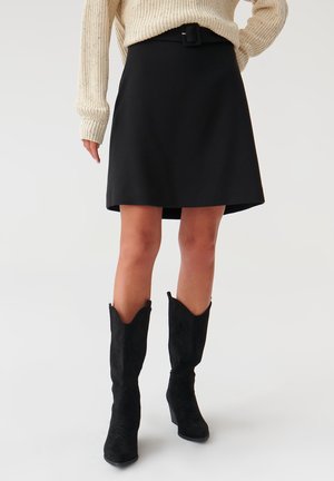 Sort A-formet nederdel af glat stof med bælte og firkantet spænde. Kombineret med tætsiddende sorte ruskindsstøvler med spidse snuder.