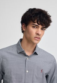 Skjorta med knäppning fram i svartvitt rutigt mönster, med spetsig krage och liten röd logotyp på vänster bröst. Bomullsmaterial.