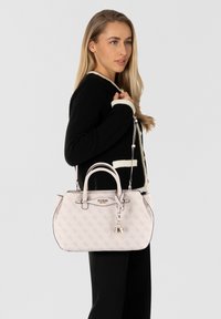 Sac à main rose pâle avec un motif monogramme, forme structurée, double anses supérieures et un charm amovible, sur un fond neutre.
