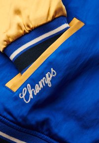 Tissu bleu avec des accents jaunes ; présente un poignet rayé et un texte brodé "Champs". Fini texturé sur l'ensemble, avec des coutures visibles.