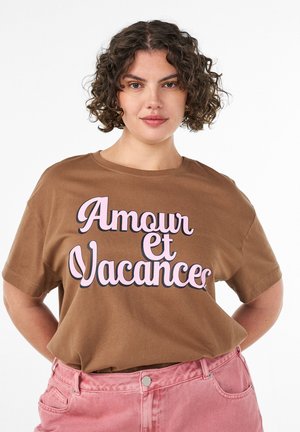Kobieta o kręconych włosach nosi brązowy t-shirt z różowym napisem „Amour et Vacances”, wpuszczony w różowe spodnie z wysokim stanem, pozując na białym tle.