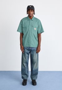 Jauns vīrietis, kurš valkā melnu Adidas cepuri, zaļu īsroku pogājamu kreklu, brīvas piegriezuma zilas džinsas un melnus apavus, stāv uz zila grīdas seguma.