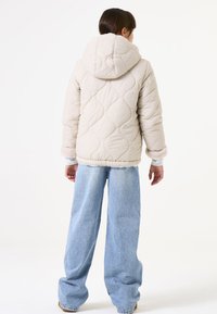 Chaqueta acolchada de color beige claro con capucha, puños forrados de piel y dobladillo. Combinada con unos vaqueros de pierna ancha azules, vista desde atrás.