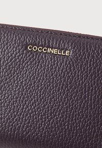 Texturovaná tmavě fialová kožená peněženka se zlatým brandingem "Coccinelle". Má drsný povrch a precizní švy. Odolný materiál.
