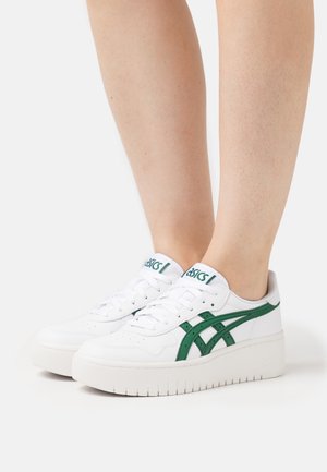 White sneakers with green accents, textured leather, flat soles, and a logo on the tongue. Rounded toe and lace-up design.