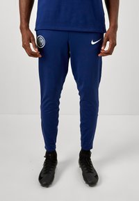 Nike bukser i marineblå polyester, tætsiddende pasform med hvide logo-detaljer; glat tekstur, elastisk talje, velegnet til sport eller casual brug.