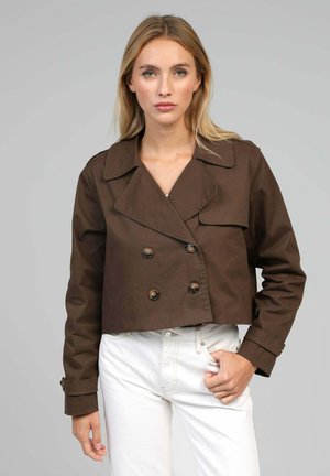 Femme blonde portant une veste courte marron à double boutonnage et un jean blanc taille haute, debout devant un fond gris.