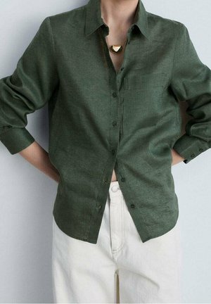 Person trägt ein dunkelgrünes Hemd mit aufgekrempelten Ärmeln, helle Hosen und eine goldene Halskette mit geometrischem Anhänger.