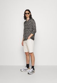 Chemise à manches longues à motif floral noir, shorts beige clair, chaussures de course argent et blanc avec des chaussettes noires ; mannequin portant des lunettes de soleil.