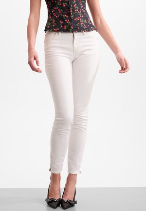 Vrouw draagt witte slim-fit broek met ritsen bij de enkels, zwarte puntige hakken met strikjes en een zwart mouwloos top met kersenpatroon.