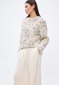 Pletený vlněný svetr v krémové barvě se šedými a béžovými skvrnami, žebrovaná textura a volný střih. Doplněno světle béžovými kalhotami na stažení.