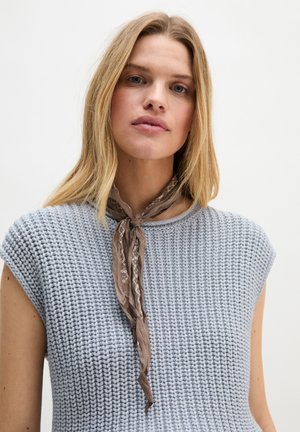 Femme blonde portant un débardeur tricoté bleu clair et un foulard marron à motif noué autour du cou, fond neutre.