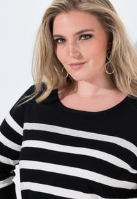 Czarny i biały sweter w paski z okrągłym dekoltem oraz srebrnym paskiem akcentującym. Modelka ma długie blond włosy i srebrne kolczyki w kształcie kół.