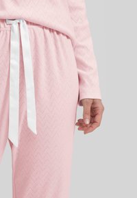 Conjunto de pijama rosa claro con textura de espiga, con un lazo de satén blanco en la cintura y mangas largas. Diseño suave y de corte relajado.