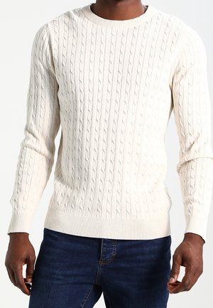 Homme portant un pull en maille torsadée crème et un jean bleu foncé, debout les bras détendus le long du corps, devant un fond uni.