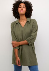 Tunic verde oliva dal taglio morbido con colletto a punta, maniche a tre quarti, tessuto texture e silhouette ampia. Abbinato a jeans blu.