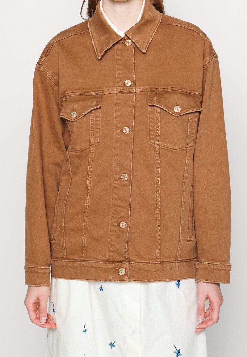 Veste en denim marron avec un col, des fermetures à boutons et deux poches sur la poitrine. Le tissu a une texture robuste et des détails de couture visibles.