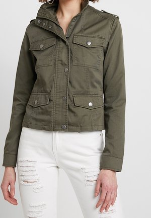 Kvinde iført en olivengrøn jakke med knapper og fire forlommer over revnede hvide jeans, stående mod en ensfarvet baggrund.