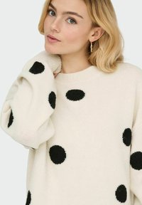 Pull oversize crème avec de grands pois noirs en feutre. Encolure et poignets côtelés ajoutant de la texture au tissu en maille.